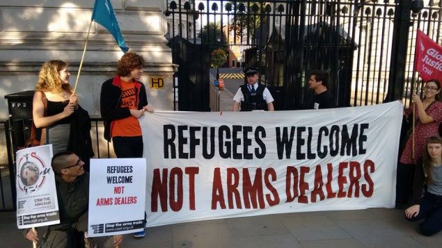 Five people stand with a banner that reads "Refugees Welcome, Not Arms Dealers"