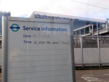 In the foreground a DLR Service Information sign says time to stop the arms fair; in the background the ExCeL centre