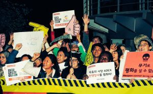 A crowd of international activists stands outside a building in the dark behind a yellow banner with hazard tape marking at the top and writing that says ADEX in English plus more in Korean. They are smiling and several of them have their hands raised in the air. One is holding a scythe. They are holding placards written Korean, Arabic and English. The English writing that can be fully seen says #stopADEX and War Starts Here.