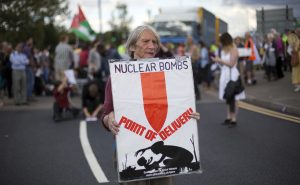 A person stands in the foreground holding a placard reading 'nuclear bombs point of delivery'. There is a red silhouette of a bomb pointing down at two figures silhouetted in black. An adult figure is crouched over a child. Also silhouetted in the background are a tank and two dead trees. The bottom of the banner reads 'Quaker Peace and Social Witness www.quakers.org.uk/qpsw'. The background of the photo is out of focus but a crowd of people can be seen standing in the road and on the pavement in front of a stationery lorry.