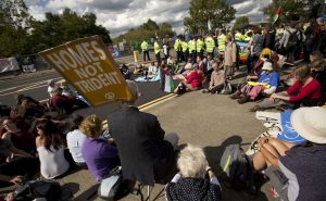 A large group of people sit in a circle in the road. Behind them there are more people standing up and a line of police in flourescent yellow jackets. Some are holding Palestinian flags and in the foreground is a brown placard bearing the CND symbol and reading 'homes not Trident'. On a fence at the other side of the road many different banners have been hung.