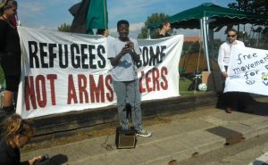 A black person with short hair, a grey t shirt and grey jeans speaks into a microphone in front of a banner which reads "Refugees welcome not arms dealers"