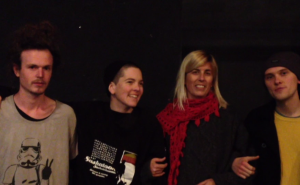 Four people facing the camera link arms in a darkened room