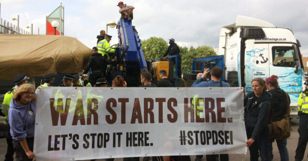 Two people hold a banner that says, War Starts Here Let's Stop It Here #StopDSEI in front of a truck carrying equpiment which is stopped in the road. Someone is sat on top of the equipment.