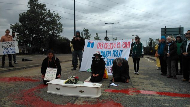 Three people kneel behind a white coffin with red paint on the road