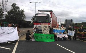 Activists bearing a sign saying No Faith in War block a truck going into DSEI