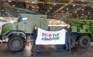 Two activists hold a 'Stop the Arms Fair' banner in front of a dark green armoured vehicle