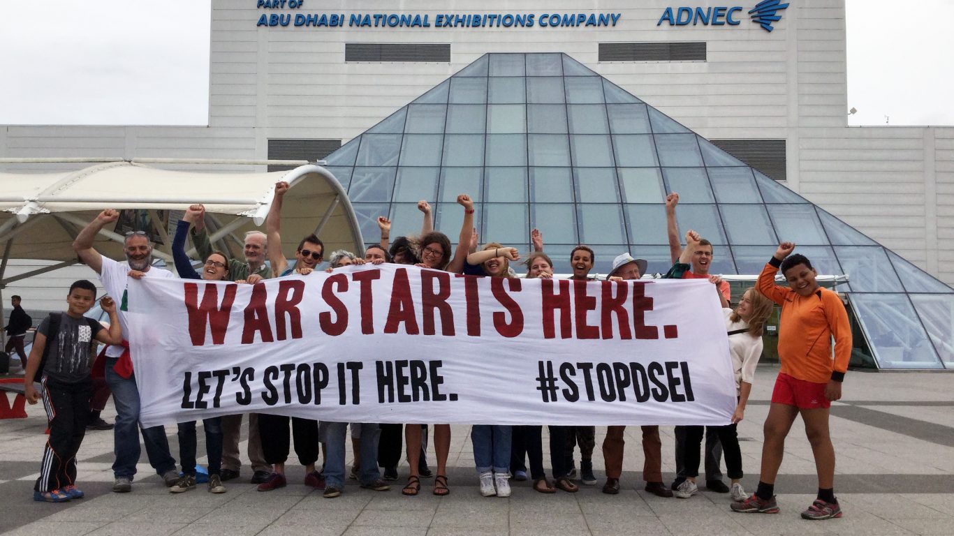 A crowd of activists hold a banner in front of the Excel centre. It says When only 0.2% of jobs in the UK are related to arms exports #stopDSEI