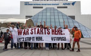 A crowd of activists hold a banner in front of the Excel centre. It says When only 0.2% of jobs in the UK are related to arms exports #stopDSEI